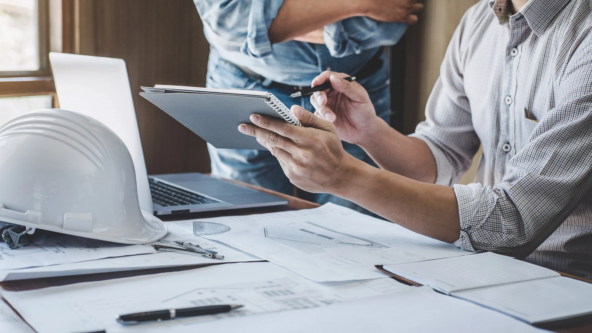 Foto von einen Mann, der an einem Schreibtisch sitzt mit mit vielen Plänen und einem Laptop darauf..
