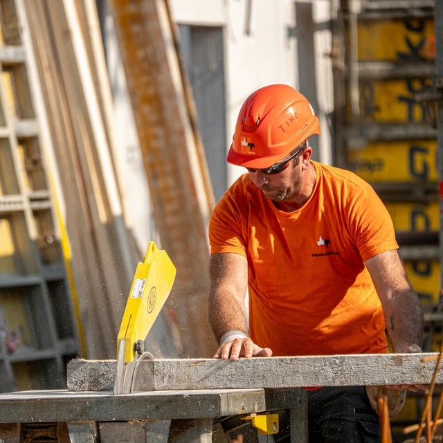 Auf einer Wilhelm-Mayer Baustelle wird fleißig gearbeitet. Ein Mitarbeiter schneidet ein Holzbrett an einer Kreissäge zu.