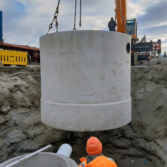 Arbeiter steht in einer tiefen Baugrube. Ein Kanalschacht wird gerade mittels Kran hineingehoben.